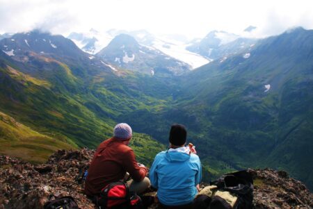 two people with hiking gear sitting on a scenic highpoint viewing Alaskan mountains